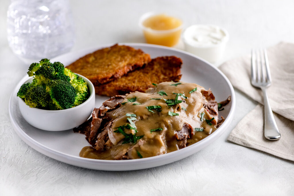 Beef brisket with brown gravy, side of broccoli and latkes with sour cream and apple sauce on a white plate.