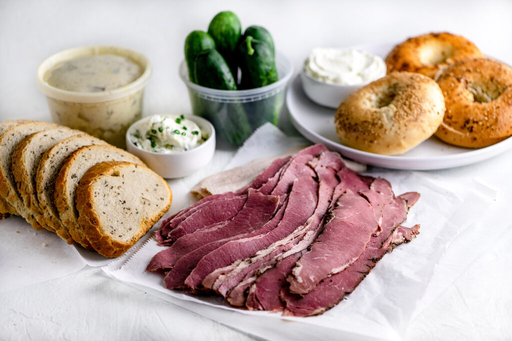 Sliced deli pastrami, corned beef and roast turkey beside a loaf of sliced rye bread, potato salad, NY bagels, cream cheese and pickles