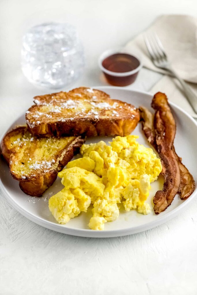 Challah french toast on white plate with scrambled eggs, bacon and syrup