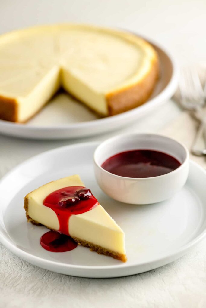 new york style cheesecake with strawberry topping on white plate with bowl of strawberry topping