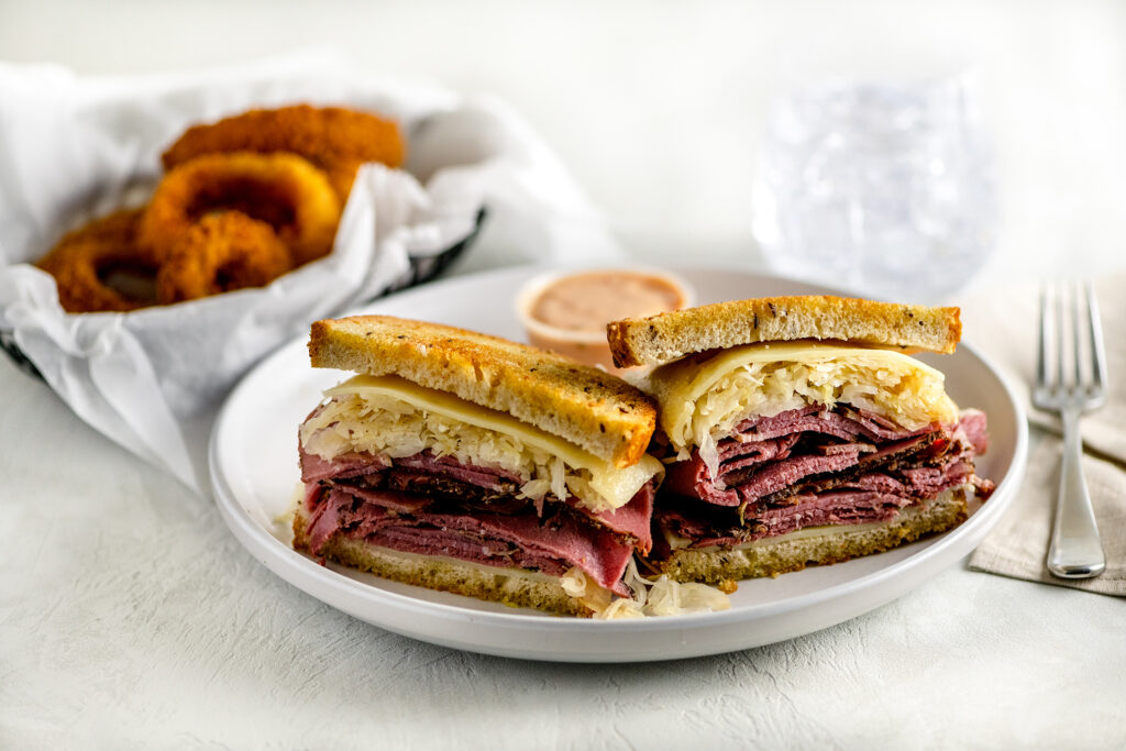 NY pastrami reuben with sauerkraut on rye bread and melted swiss cheese on a white plate. Large order of jumbo crispy onion rings as a side. 