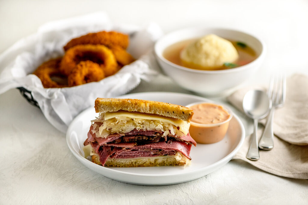 New York Pastrami Reuben on a white plate with onion rings and matzo ball soup