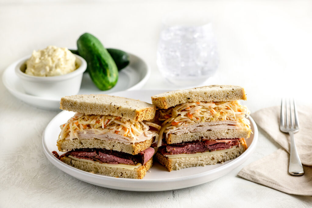 triple decker sandwich with pastrami, turkey and cole slaw between 3 slices of rye bread on a white plate. Pickles and potato salad on the side.