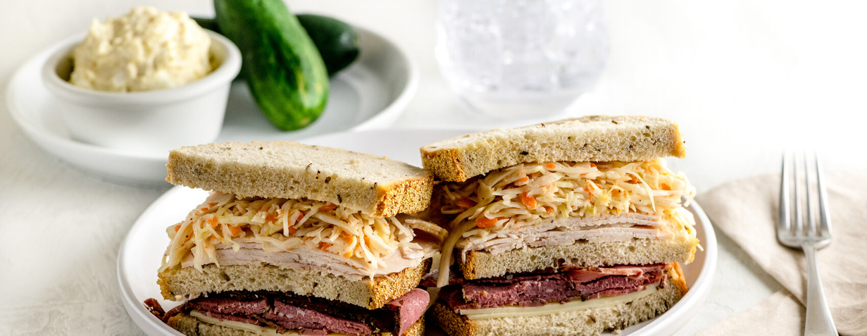 triple decker sandwich with pastrami, turkey and cole slaw between 3 slices of rye bread on a white plate. Pickles and potato salad on the side.