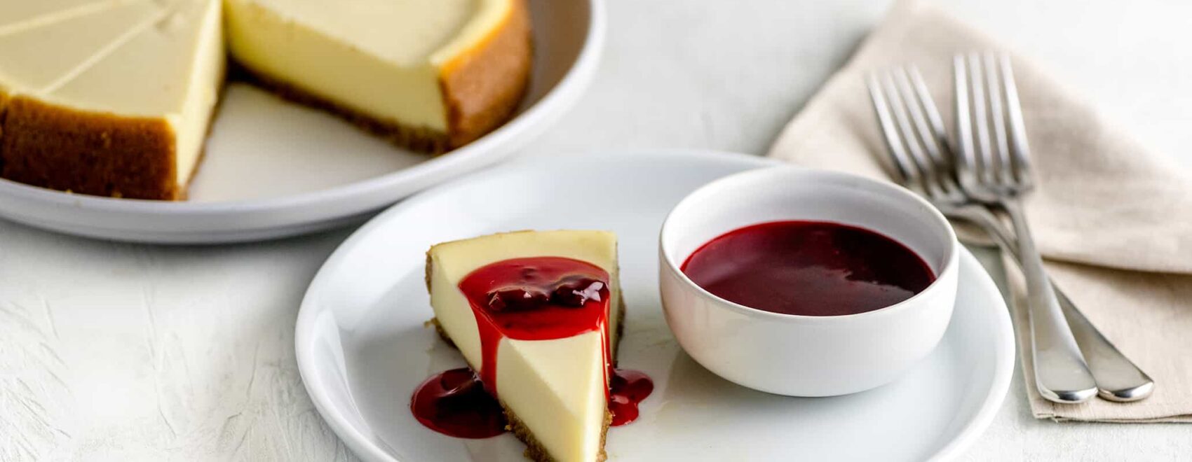 Homemade cheesecake on a white plate beside a small bowl of strawberry topping