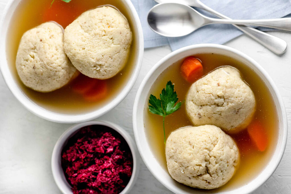 Two white bowls of matzo ball soup in broth with carrots