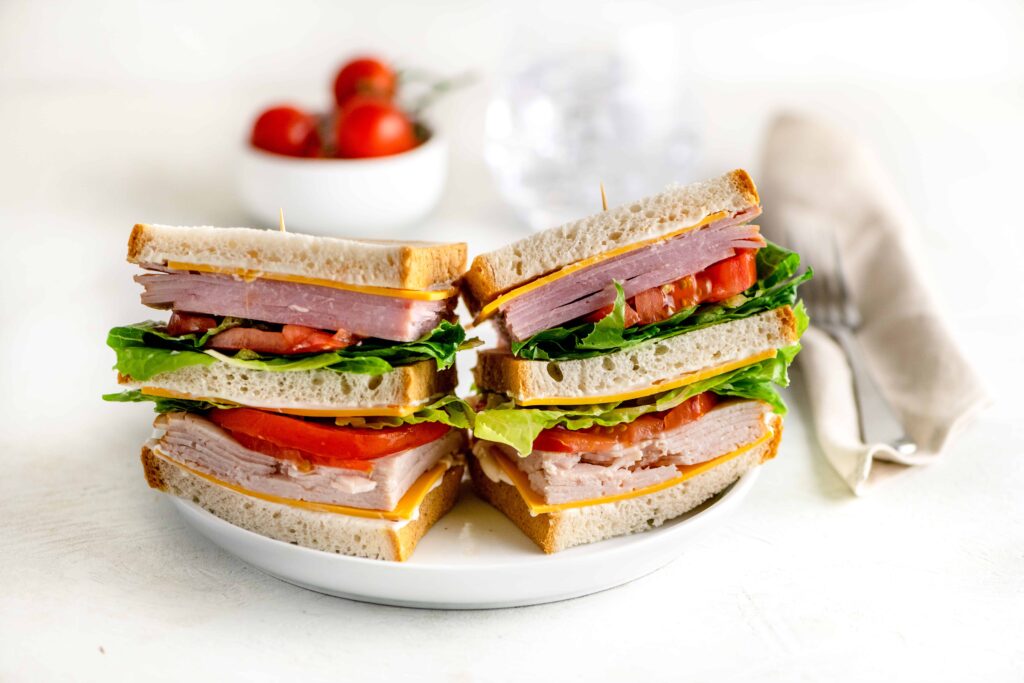 Triple decker sandwich of ham, roasted turkey on three pieces of gluten free bread with cheese, lettuce, tomato and mayo on a white plate