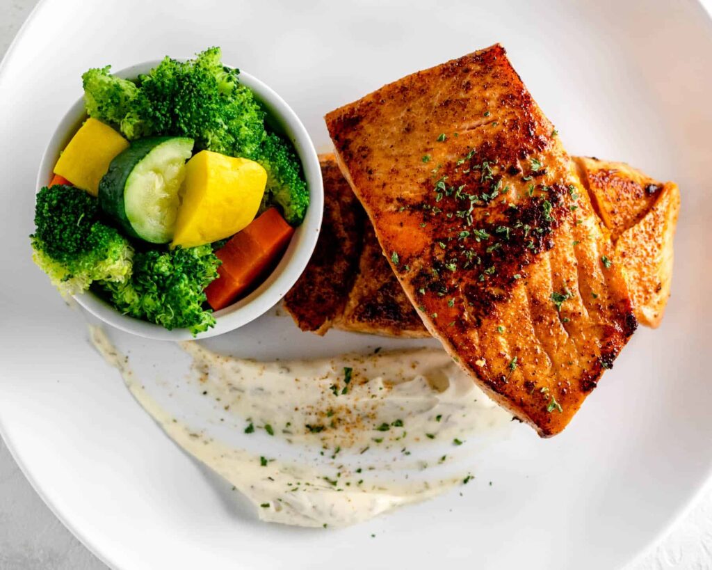 two grilled salmon fillets with steamed veggies and dill mayo on a white plate