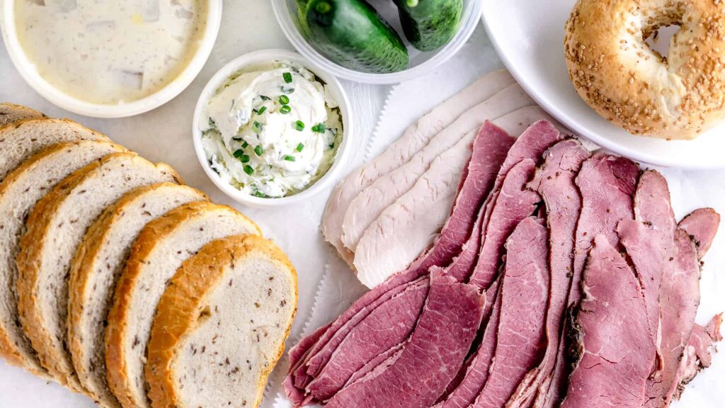 Spread of Jewish Rye bread, chive cream cheese, sliced corned beef, pastrami, turkey and New York bagels on a white table top.