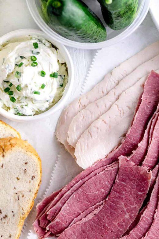Spread of Jewish Rye bread, chive cream cheese, sliced corned beef, pastrami, turkey and New York bagels on a white table top.